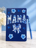 Navy A5 notebook with large 'MAMA' floral bow and butterflies; blue pen beside it