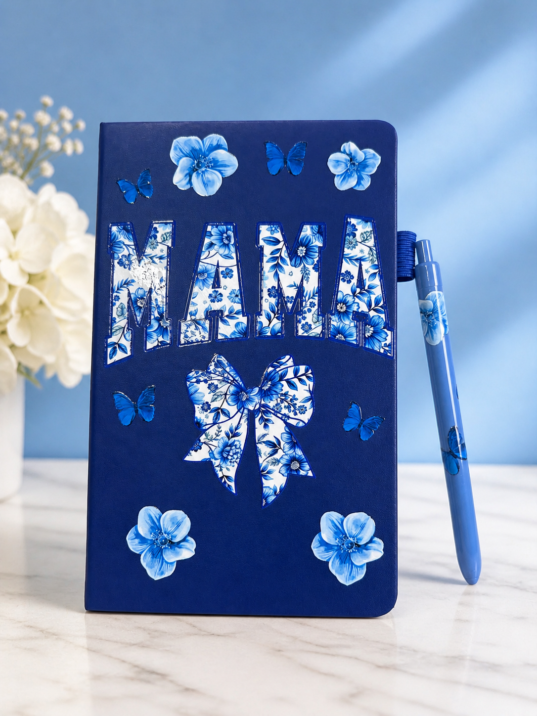 Navy A5 notebook with large 'MAMA' floral bow and butterflies; blue pen beside it